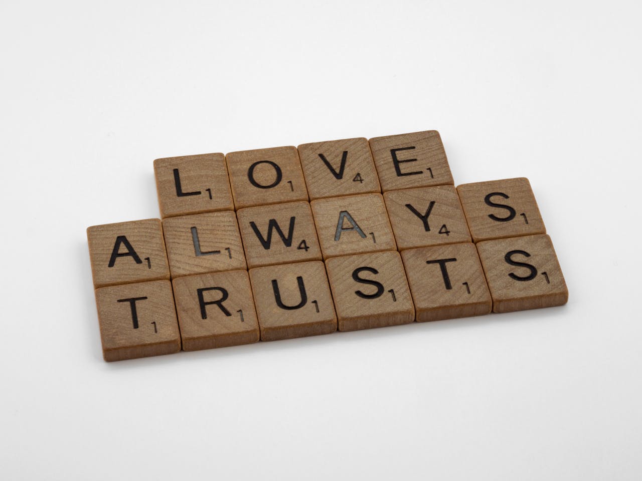 Close-up of wooden Scrabble tiles spelling 'Love Always Trusts' on white surface.